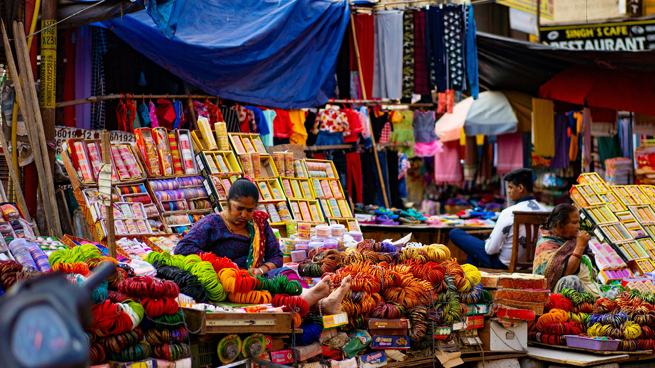 Marknad i Indien. Färgstarka föremål och en kvinna som sitter mitt bland dem.