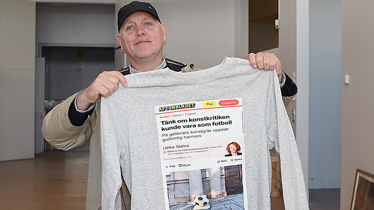 En man i keps håller upp en grå tröja med ett tryckt tidningsurklipp.